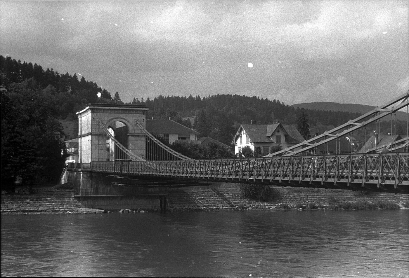 Historische Schwarzweissaufnahme der alten Aarauer Kettenbrücke: eine schmale Hängebrücke über die Aare, deren Ketten über einen wuchtigen Steinturm mit Rundbogendurchfahrt laufen, im Hintergrund bewaldete Hügel