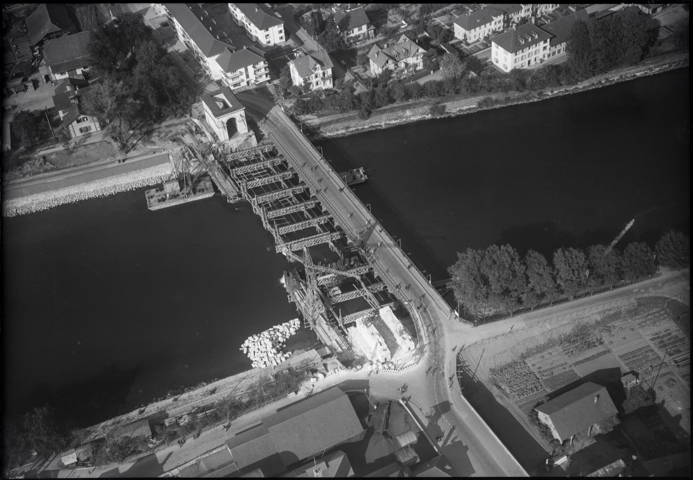 Schwarzweisses Luftbild der Baustelle: neben der bestehenden Brücke steht ein hölzernes Lehrgerüst im Fluss, am linken Ufer der noch stehende steinerne Torturm, ringsum Wohnhäuser und Gärten