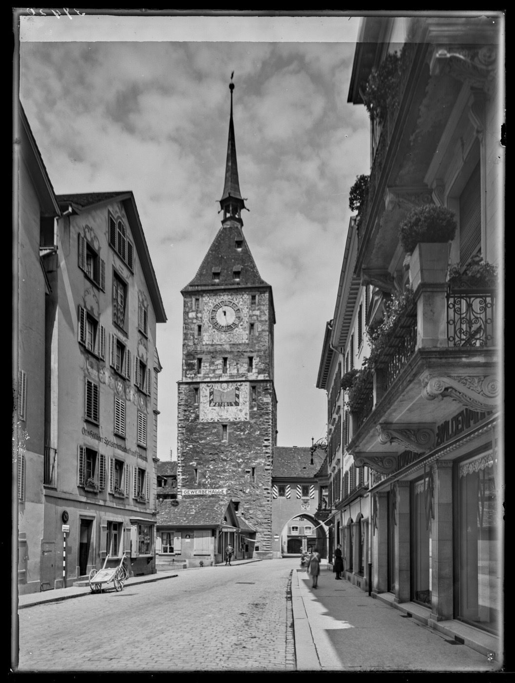 Historische Schwarzweissaufnahme: Blick eine Gasse hinunter auf den Oberen Turm mit grossem Uhrzifferblatt und darunter einer bemalten Sonnenuhr, links ein Haus mit verzierter Fassade, auf der Strasse ein Handwagen