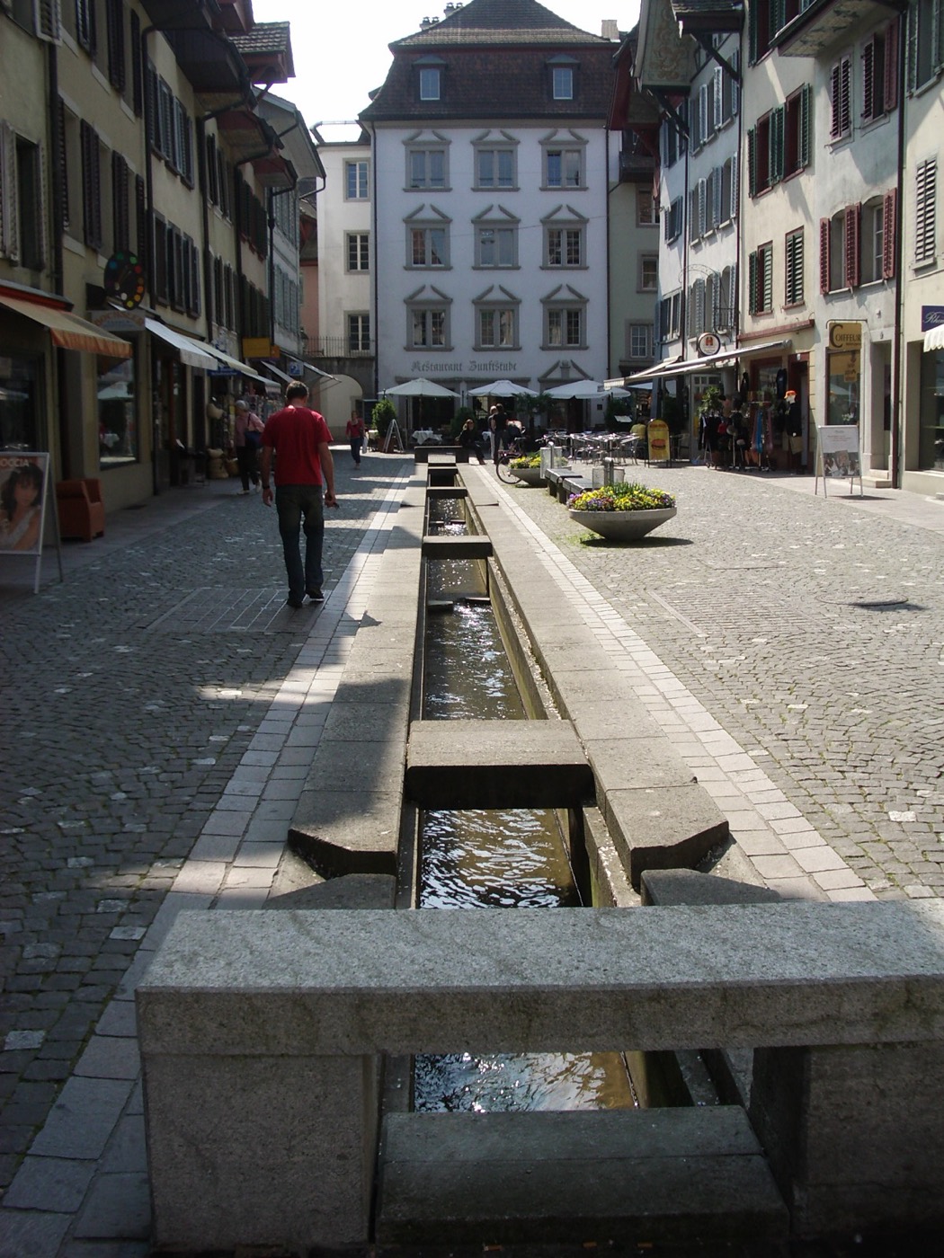 Der offene Stadtbach läuft in einem schmalen Steinkanal mitten durch eine Aarauer Altstadtgasse, überquert von flachen Steinplatten, links und rechts Häuserzeilen mit Läden