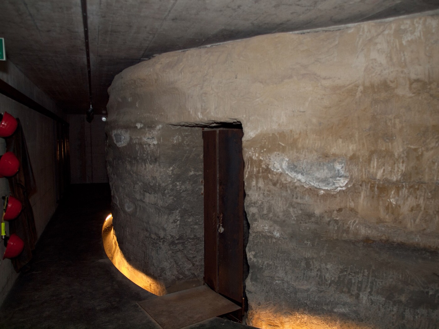Blick in einen Meyerschen Stollen: ein aus dem Fels gehauener, gemauerter Gang mit rundem Gewölbe und einer schmalen Eisentür, an der linken Wand hängen rote Schutzhelme